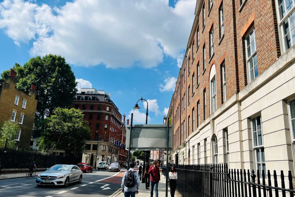 Pedestrians walking along a brick-fronted London street with railings, trees and a cycle lane